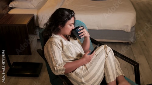 woman scrolling on phone at night, she is seating on couch and drinking coffee while watching reels in phone