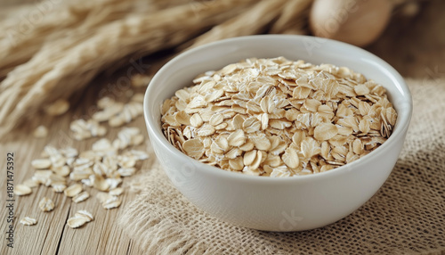 White bowl filled with rolled oats sits on rustic burlap. Close up shows raw cereal flakes, perfect for healthy breakfast or snack