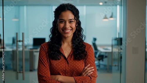Confident Hispanic businesswoman walking forward and crossing arms with a bright smile in a modern glass office, illustrating professional success and female leadership.