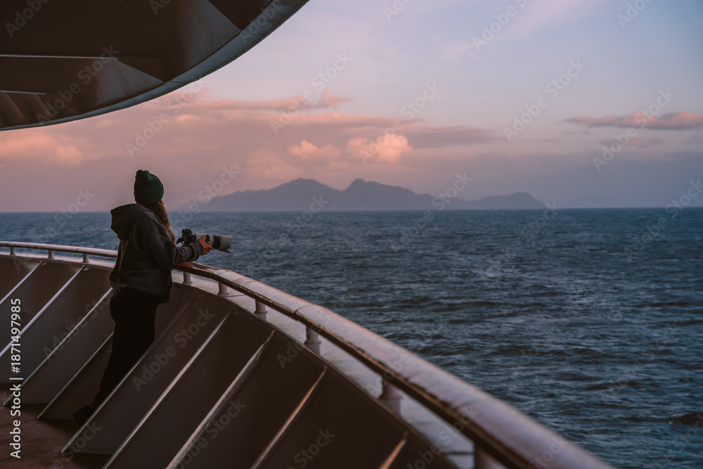 Fototapeta premium Female Photographer on Cruise Ship Looks At Mountain at Sunset. Adventure Travel. Beautiful Girl On Deck Handrail Balcony Looks Over Ocean