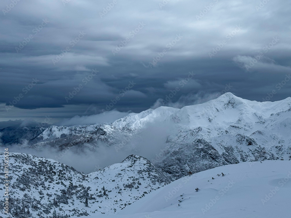 Obraz premium Dark winter storm clouds hang over a rugged mountain range covered with fresh snow. Mist rolls through the valleys, adding to the whimsical alpine atmosphere. Dramatic views in snowy Slovenian Alps.