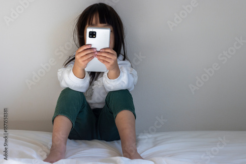 うす暗い部屋で座ってスマートフォンを見る女の子のイメージ（5歳、日本人、顔無し）