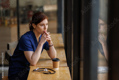 Fotografie A female nurse in blue scrubs appears troubled while holding a coffee cup outside a medical facility, reflecting the stress of healthcare professionals