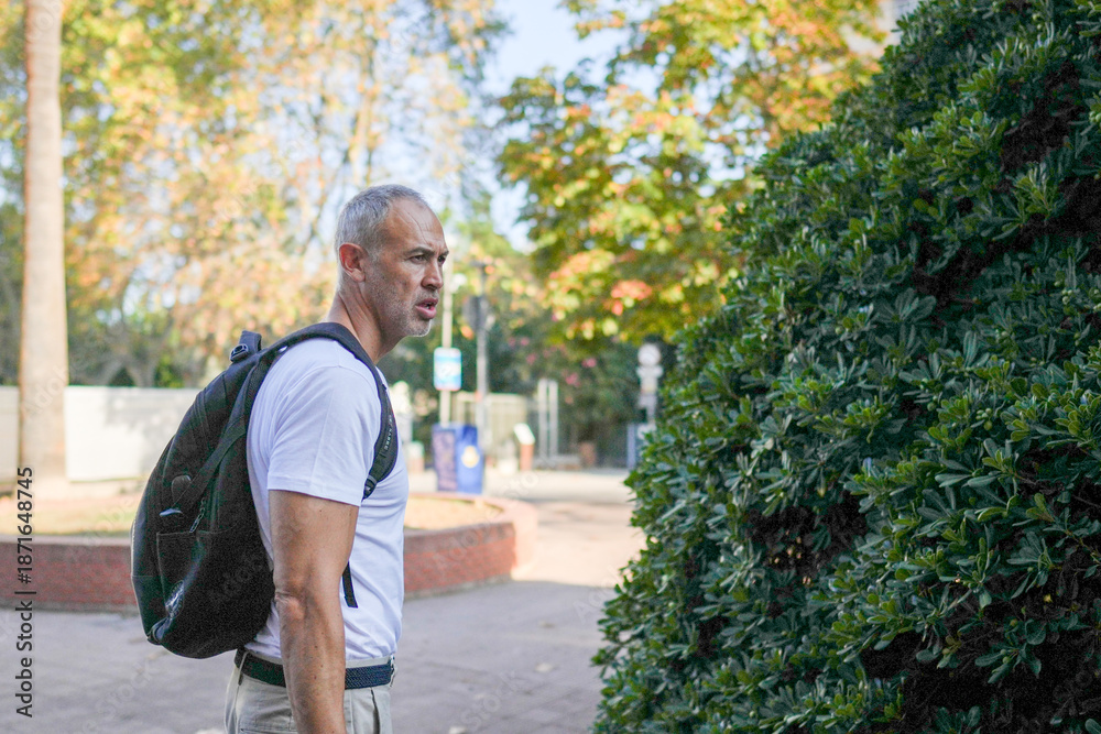 Obraz premium A man wearing a backpack is standing in front of a bush. He looks like he is lost or confused