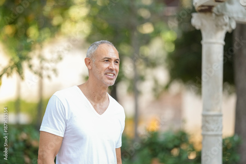 A man in a white shirt is smiling and looking at the camera. He is enjoying himself and is in a park setting