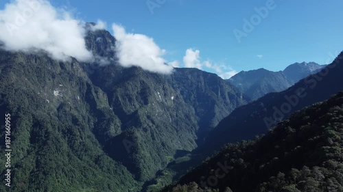 Aerial view of lush green mountain valley with drifting clouds under clear blue sky, creating a peaceful tropical highland landscape for nature and travel themes.