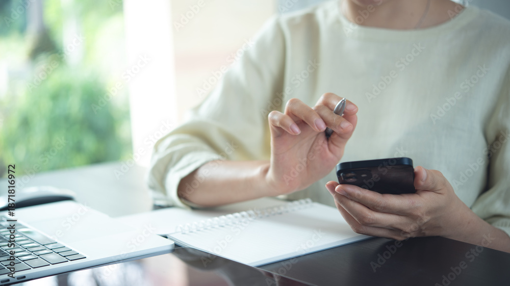 Fototapeta premium Close-up of a professional holding a smartphone and pen beside a laptop and notebook, showing multitasking, planning, and modern remote work.