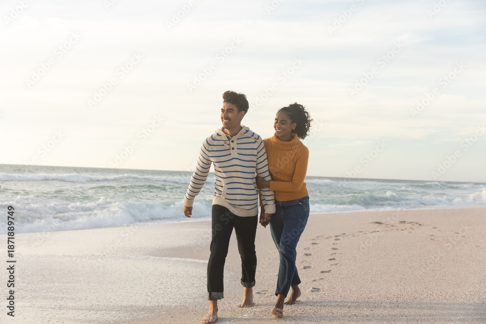 Fototapeta premium Full length of happy multiracial couple walking on shore at beach against sky during sunset