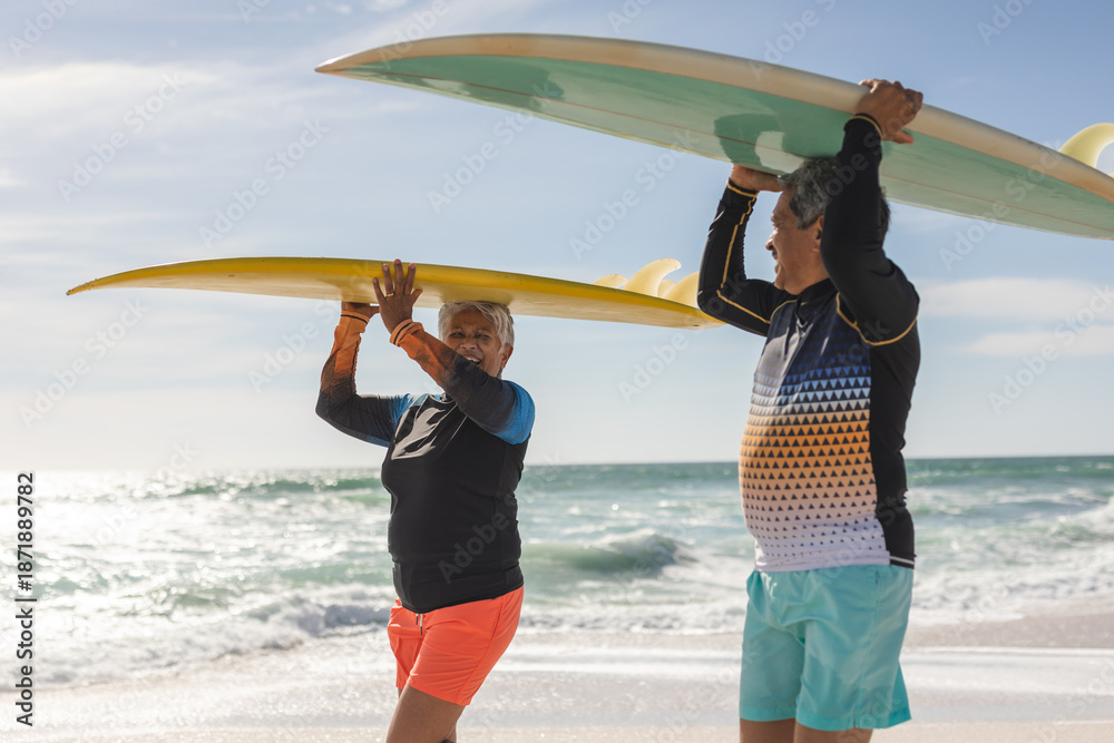 Fototapeta premium Happy multiracial senior couple looking at each other while carrying surfboards on heads at beach