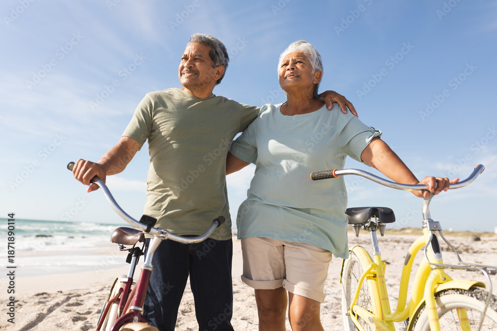 Fototapeta premium Smiling senior multiracial couple looking away with bikes at beach against sky during sunny day