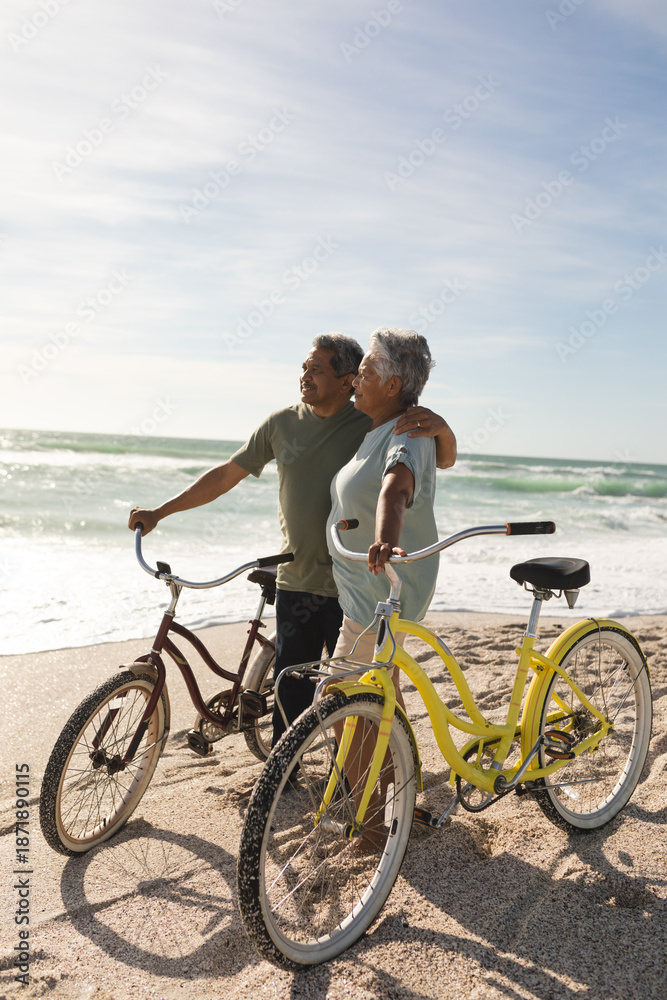 Fototapeta premium Multiracial senior couple looking away standing with bicycles at beach against sky during sunny day