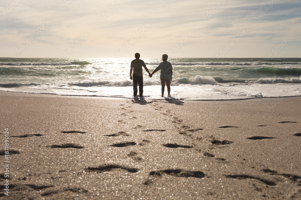 Fototapeta premium Footprints on sand with multiracial senior couple holding hands standing at beach during sunny day