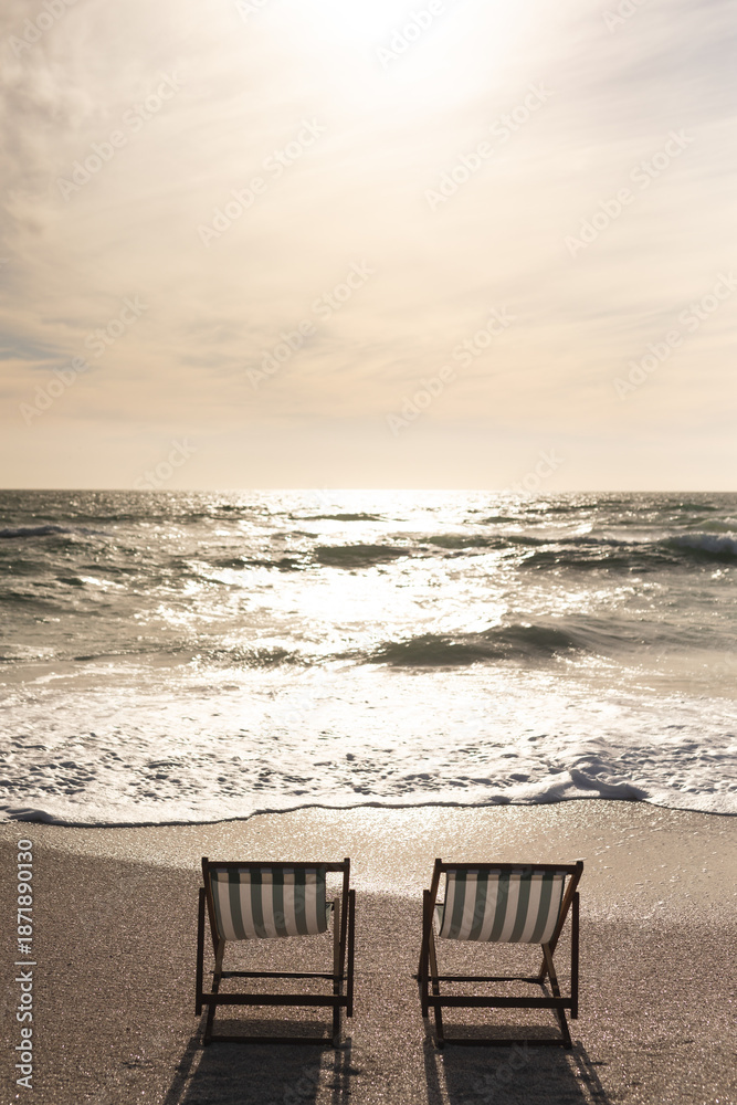 Fototapeta premium Two empty folding wooden chairs on shore in front of waves splashing in sea at beach against sky