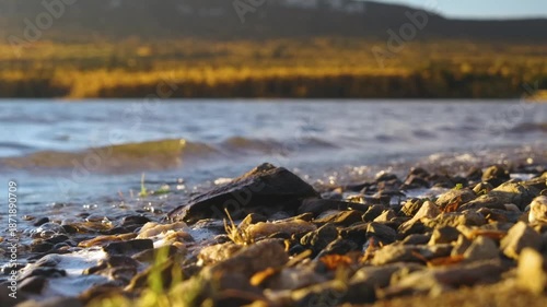 Close-up shot of the rocky shore of a lake or river in the Southern Urals region of Russia. The golden light of the setting or rising sun illuminates the pebbles and gentle waves, creating a peaceful,