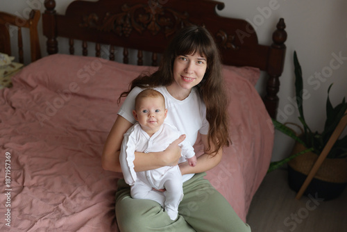 Mother sits on bed with baby, both smiling. The room has soft light and a simple, warm atmosphere. They seem to enjoy their time together