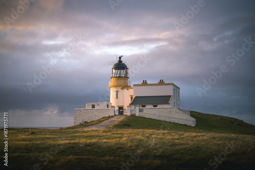 View of a gleaming white lighthouse perched atop a grassy hill under a dramatic sky of swirling blues and oranges, Isle of Skye, United Kingdom.