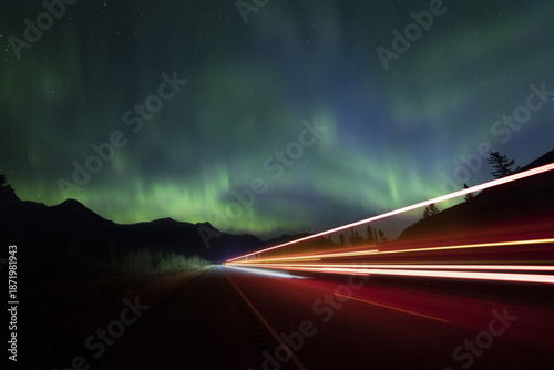 View of vibrant aurora borealis dances above the dark silhouettes of mountains, as car lights streak across the highway, Jasper, Alberta, Canada.