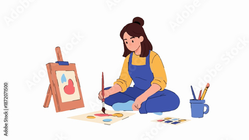 A person in a blue jumpsuit and yellow shirt sits cross-legged, painting on a canvas and paper with brushes