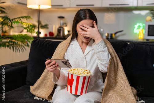 Tableau sur toile Young woman at night home watches horror on smartphone hugs blanket flinches feels fear so quick breath