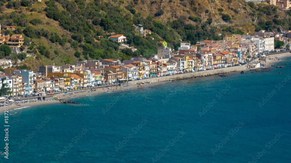 Fototapeta premium Aerial view of a vibrant coastal town nestled between a lush green hillside and the deep blue sea. It is the Giardini Naxos waterfront, located in province of Messina, Sicily, Italy. Summer morning.