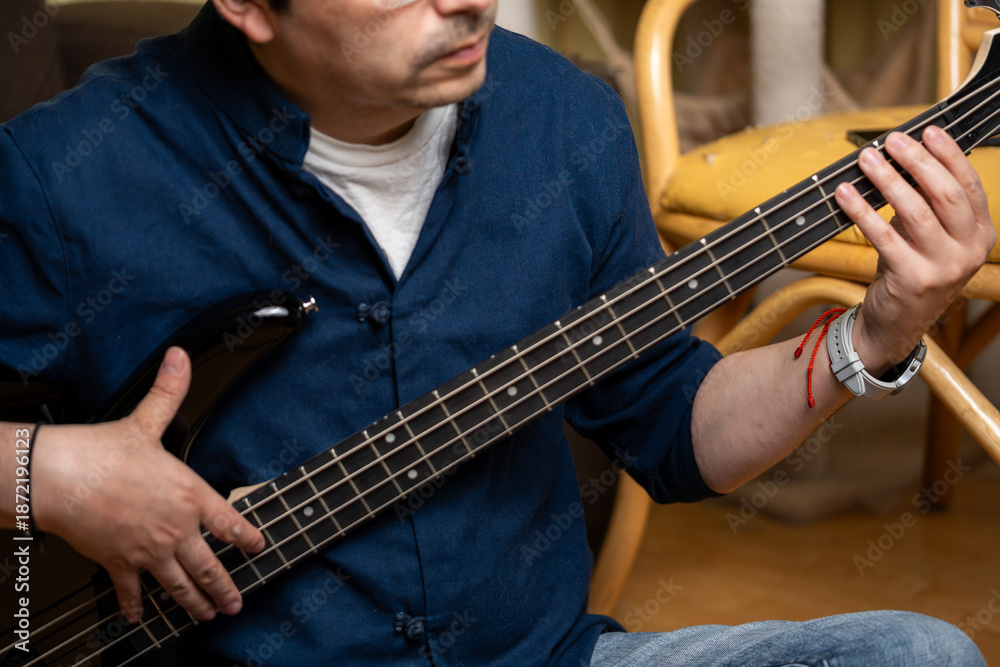 Obraz premium Middle aged man playing black electric bass guitar sitting on floor at home, Musician practicing bass guitar scales while sitting in a living room