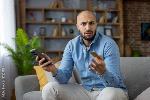 Fotografi Man looking confused and frustrated while holding a smartphone and credit card,