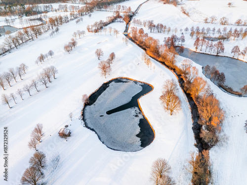 Aerial drone view of snowy golf course with frozen pond, creek and glowing sunrise trees