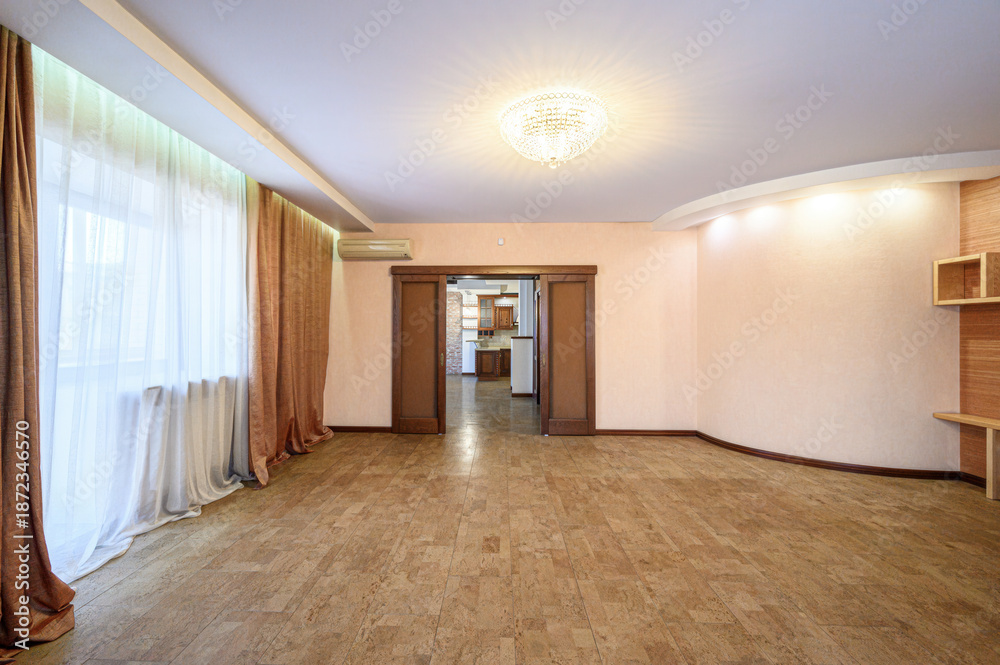 Fototapeta premium A spacious room with beige tiled flooring, large windows with sheer white and golden curtains, a crystal chandelier, wooden door frames, and built-in shelves. Light and airy