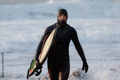 Fotografie Surfer man with surfboard in winter wetsuit walk on beach