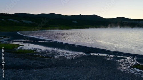 Witness the stunning beauty of the sulfatna field on Kunashir Island. Geysers bubble and steam rises against the backdrop of majestic mountains at twilight. Nature's wonder comes alive.