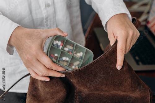 A hand holds a green, divided pill organizer filled with assorted tablets and capsules, set against a brown suede bag. The scene conveys health management, daily medications, and personal care.