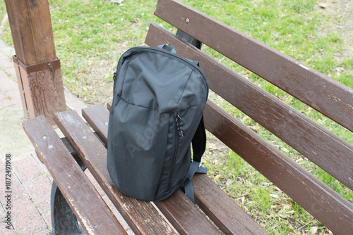 Black backpack standing on a wooden park bench in a public park. Concept of travel gear, everyday carry, lost or unattended item with copy space.