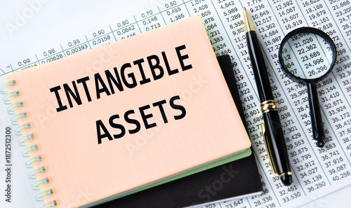 INTANGIBLE ASSETS - words on a yellow notebook on a background of digital tables, a magnifying glass and a pen