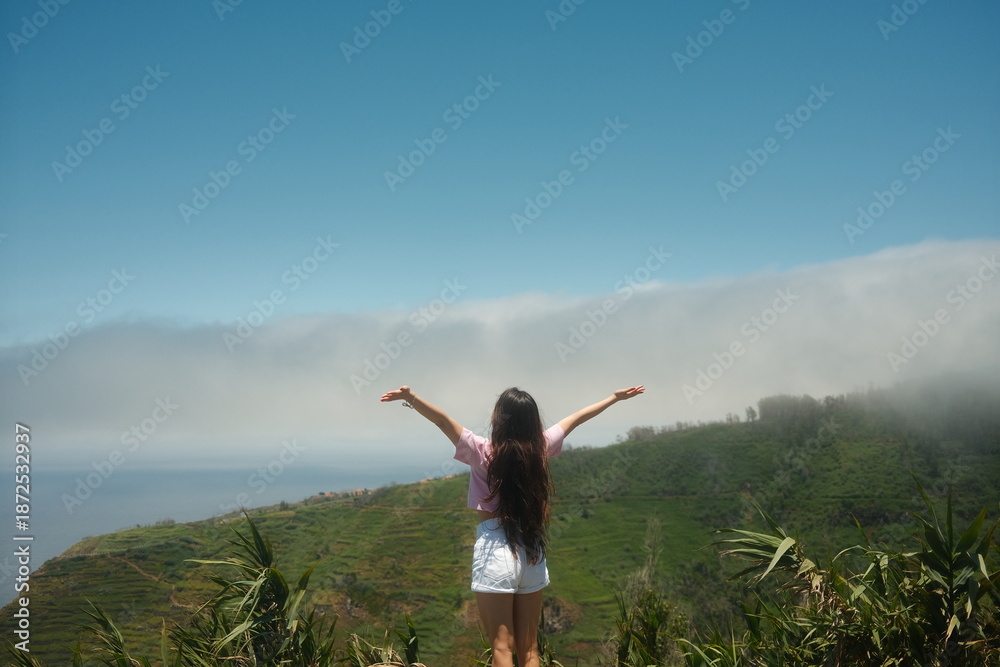 Fototapeta premium A wide shot of a young woman with arms open overlooking misty ocean landscape. Concept of solo traveling.