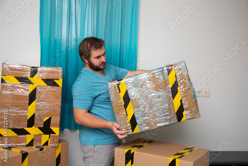 Fotografie Mature man with beard looks at picked up cardboard box with yellow and black tape in empty room of apartment