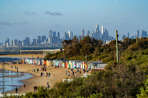 Fotografie Vibrant bathing boxes line Brighton Beach, filled with people, overlooking the calm bay and Melbourne's impressive city skyline under a clear sky