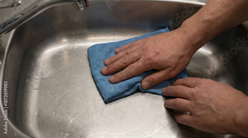Hands wiping a stainless steel kitchen sink with a blue microfiber cloth