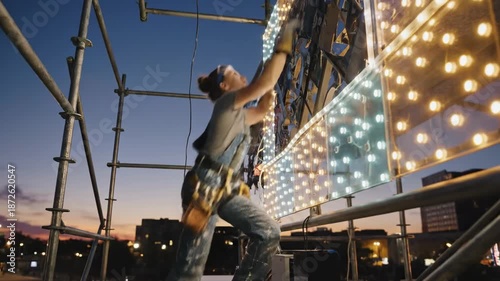 A woman in a hard hat and safety gear works on a large wall of lights on scaffolding at dusk