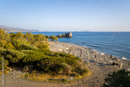 Tableau sur toile Wide view of a pebbly beach lined with closed umbrellas and lush coastal vegetation beside calm, clear blue water