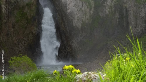 footage of the powerful Dzhily-Su waterfall cascading down a rocky cliff face in the beautiful, rugged landscape of the North Caucasus region. Lush green grass and yellow flowers are visible in the fo