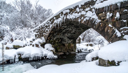An arched stone bridge covered in snow and icicles spans a flowing stream in winter