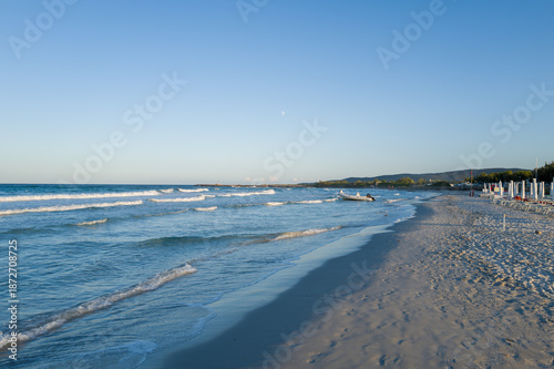 Tranquil evening view of San Teodoro beach in Sardinia, featuring soft waves lapping against smooth sand under a clear sky, with distant hills and empty sun loungers along the coast.