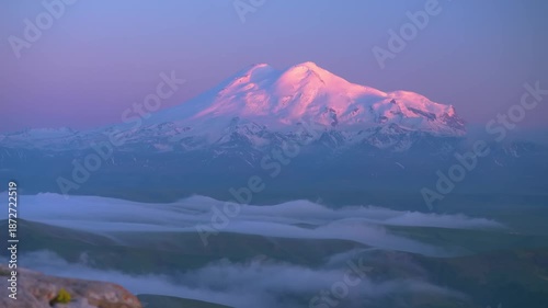 Panoramic view of the snow-capped peaks of Mount Elbrus, the highest mountain in Europe, during a vibrant sunrise or sunset, with a layer of clouds below. Located in the beautiful region of Kabardino-