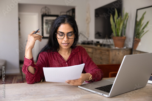 Obraz na płótnie Focused young Indian woman in glasses reading paper letter at home, analyzing fi