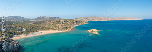 Expansive panoramic image of Vai beach in Crete, showing golden sand, lush greenery, and vivid turquoise water meeting rugged hills under a clear blue sky. The tranquil Mediterranean setting radiates