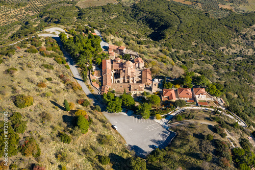 Aerial shot of the Osios Loukas Monastery complex surrounded by dense greenery and rugged hills in Central Greece, with terracotta roofs and stone walls bathed in bright sunlight. The historic site