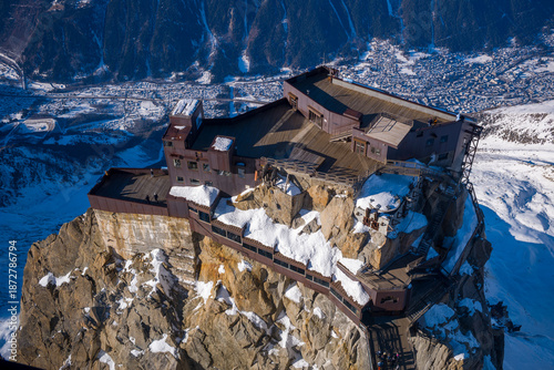 Fotografie Aerial view of the Aiguille du Midi summit station perched dramatically on rugged, snow-dusted cliffs above the Chamonix valley in bright winter sunlight
