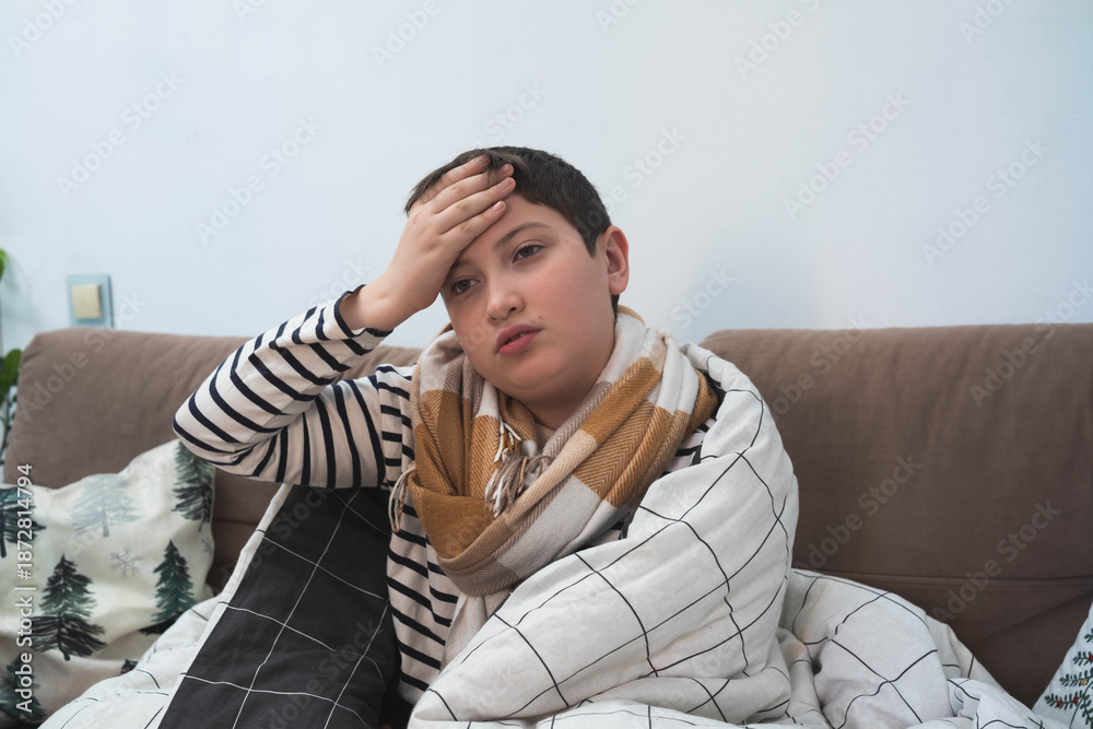 Fototapeta premium A young boy wrapped in a checkered blanket and wearing a scarf sits on a sofa. He looks unwell, suggesting he has a headache or fever. A cozy pillow with a tree pattern is beside him.
