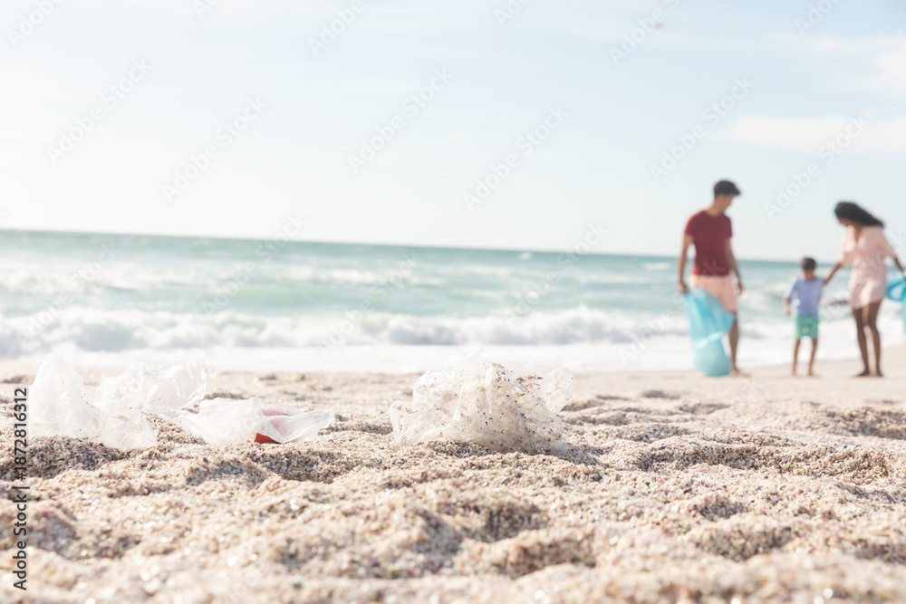 Fototapeta premium Garbage on sand with multiracial family cleaning beach on sunny day against sky with copy space