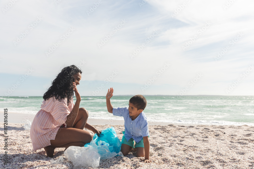 Fototapeta premium Happy mother and son giving high-five while collecting garbage in bag at beach against sky